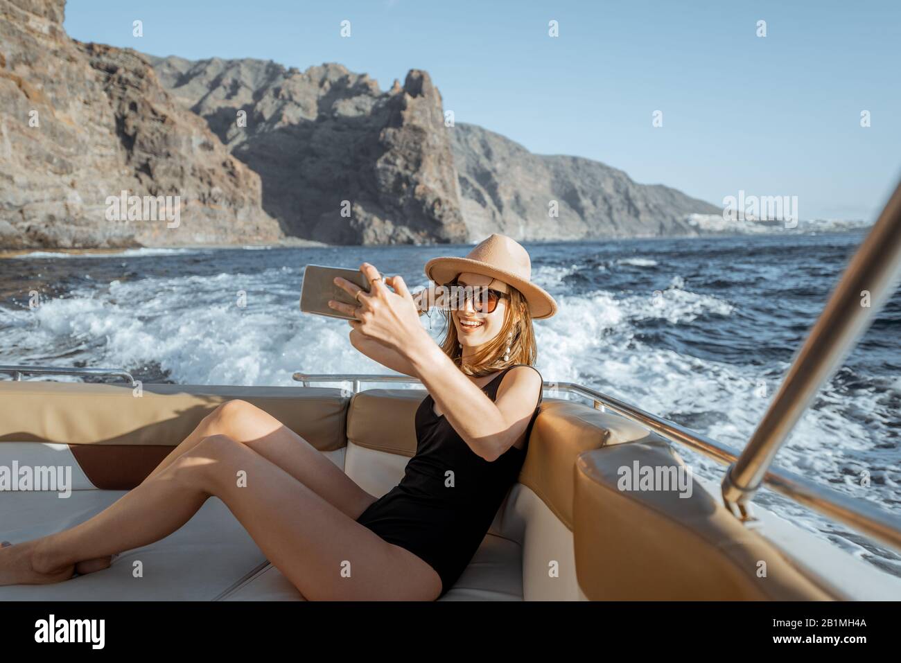 Frau in Badeanzug und Sonnenhut genießen Ozeanreise, machen Selfie-Foto am Telefon mit schönen felsigen Küste auf dem Hintergrund. Konzept einer glücklichen Sommerruhe am Meer Stockfoto