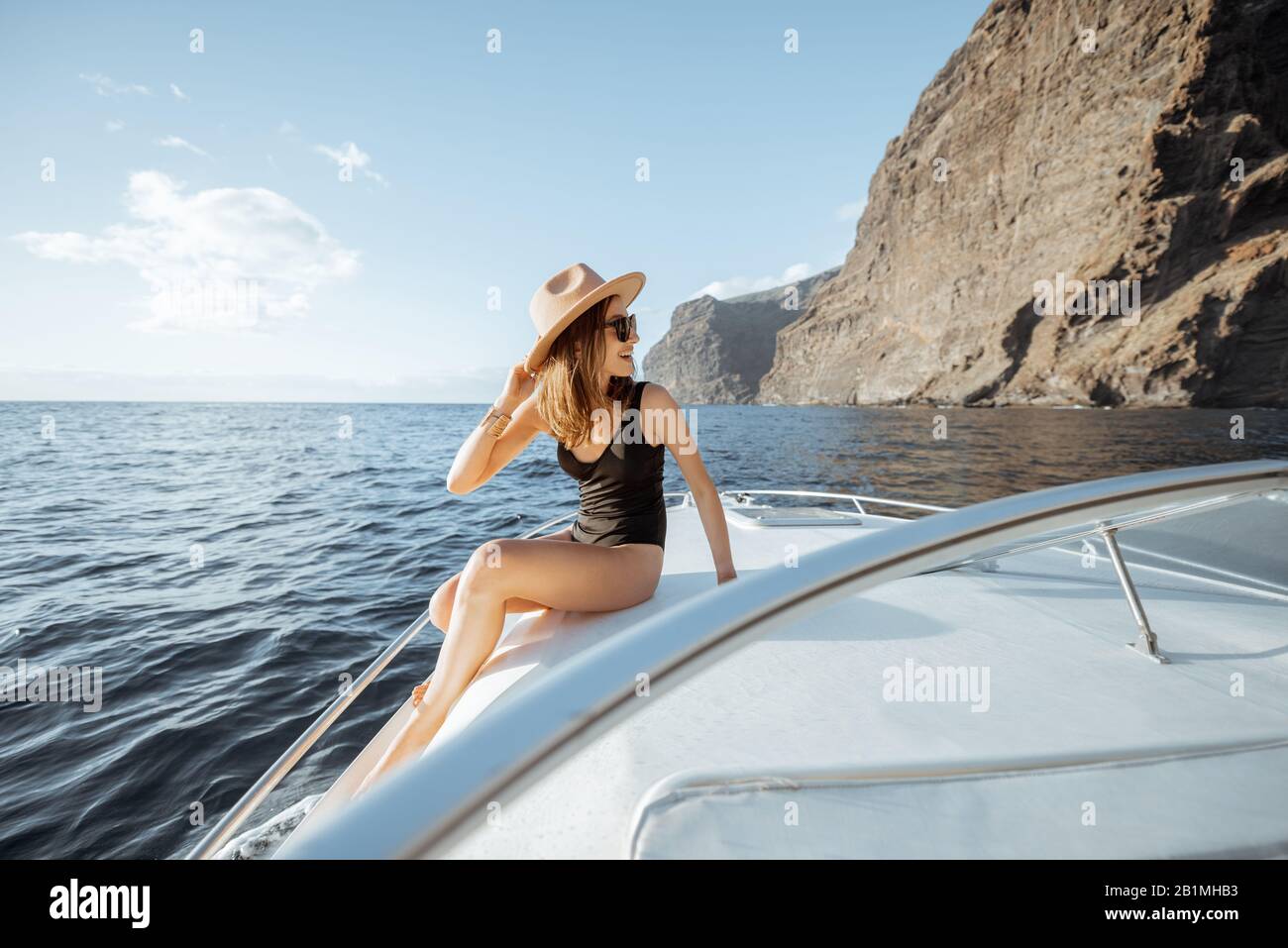 Frau in Badeanzug und Sonnenhut genießen Ozeanreise, Segeln auf einer Yacht in der Nähe der atemberaubenden felsigen Küste bei einem Sonnenuntergang. Konzept eines luxuriösen Sommer Freizeit Verfolgung und Reisen Stockfoto