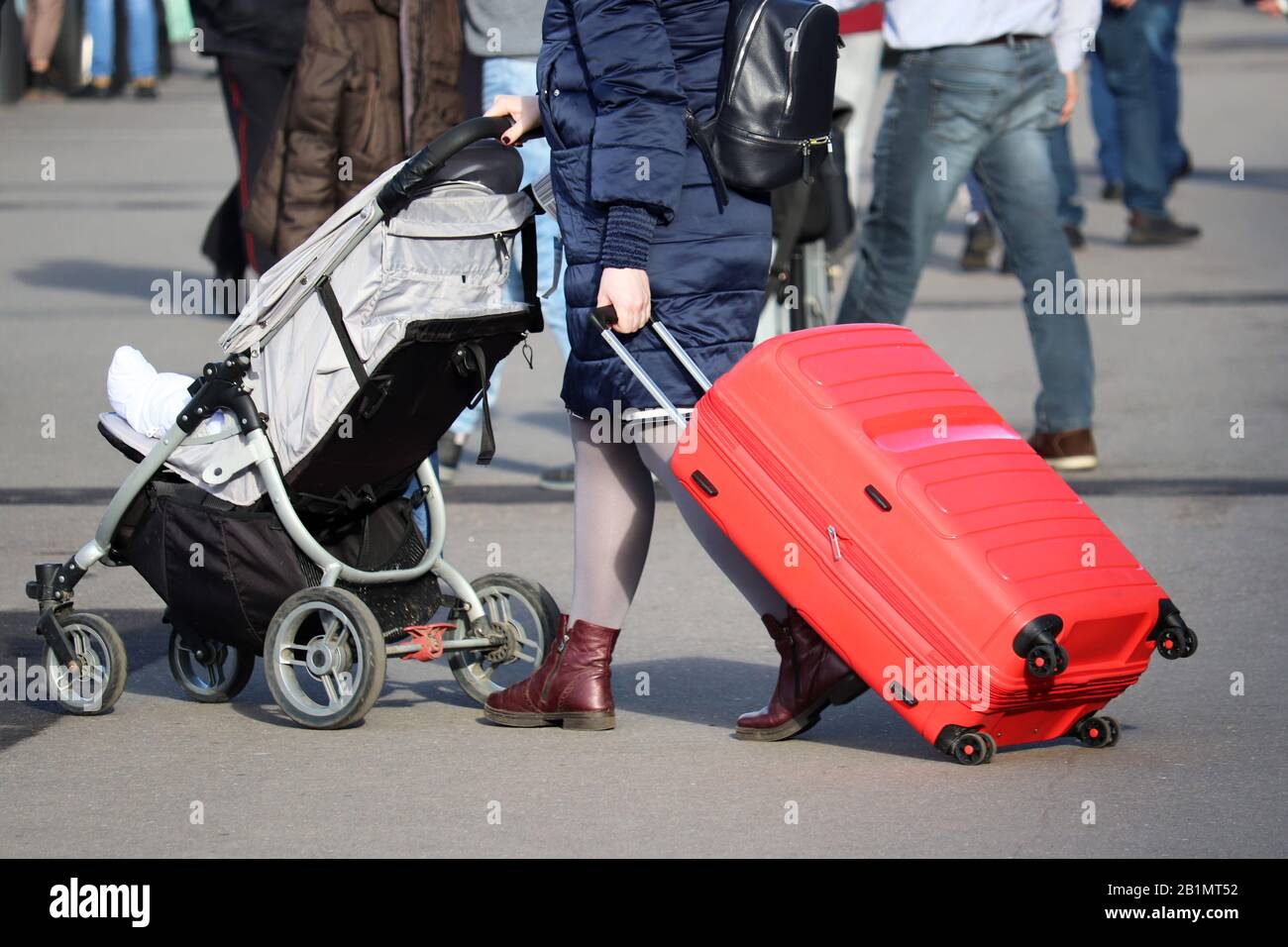 Frau, die mit einem Koffer auf Rädern und Babywagen läuft. Weibliche Beine und Gepäck auf der Straße mit Menschenmenge, Konzept der alleinerziehenden Mutter auf Reisen Stockfoto