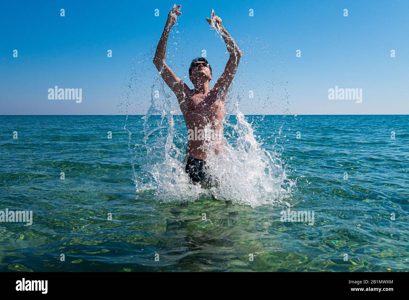 Junger Mann mit Sonnenbrille, die im Sommer aus dem Ozean springt und Spritzer verursacht, Rhodes, Griechenland. Lizenzfreies Foto. Stockfoto