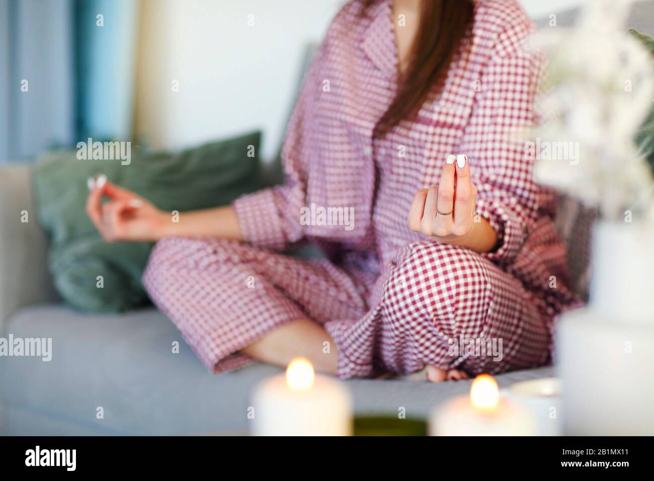 Optimistische Dame mit einer Tasse heißen Getränks und Kerzen, die im Yoga sitzt, posieren auf dem Sofa im gemütlichen Wohnzimmer. Nahaufnahme Stockfoto