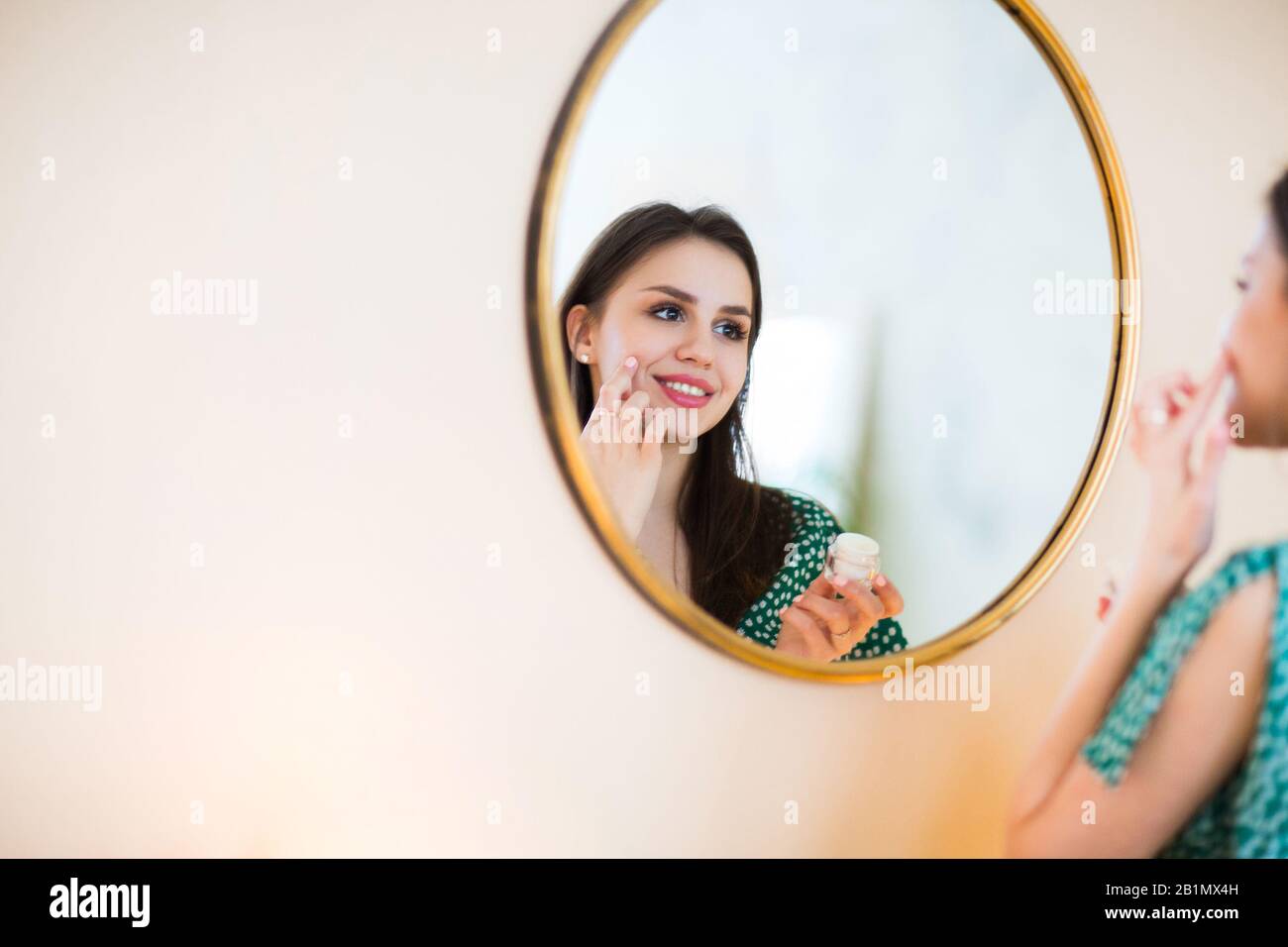 Fröhliche Brunette im grünen Kleid mit Punktmuster, feuchtigkeitsspendende Gesichtshaut mit Creme auf Kamera2 Stockfoto