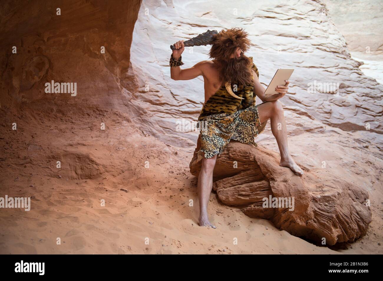 Steinzeit luddite Caveman kratzt sich mit einem Club den Kopf, während er seine Steintablette draußen in einer verwitterten Felshöhle betrachtet Stockfoto