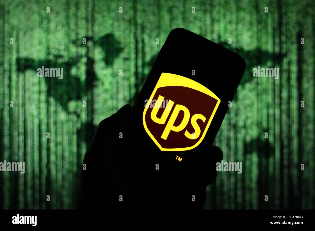 Das UPS-Logo der Versandfirma wird auf dem Smartphone-Bildschirm mit einer Matrix-ähnlichen Weltkarte angezeigt, die im Hintergrund verschwommen zu sehen ist Stockfoto