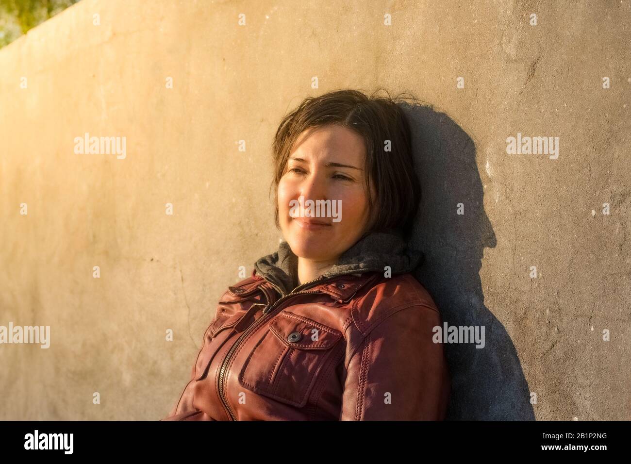 Brünette Frau mit roter Lederjacke, die sich an eine graue Betonwand lehnt, die allein unter dem Sonnenlicht sitzt. Traurigkeit und Einsamkeitskonzept. Stockfoto