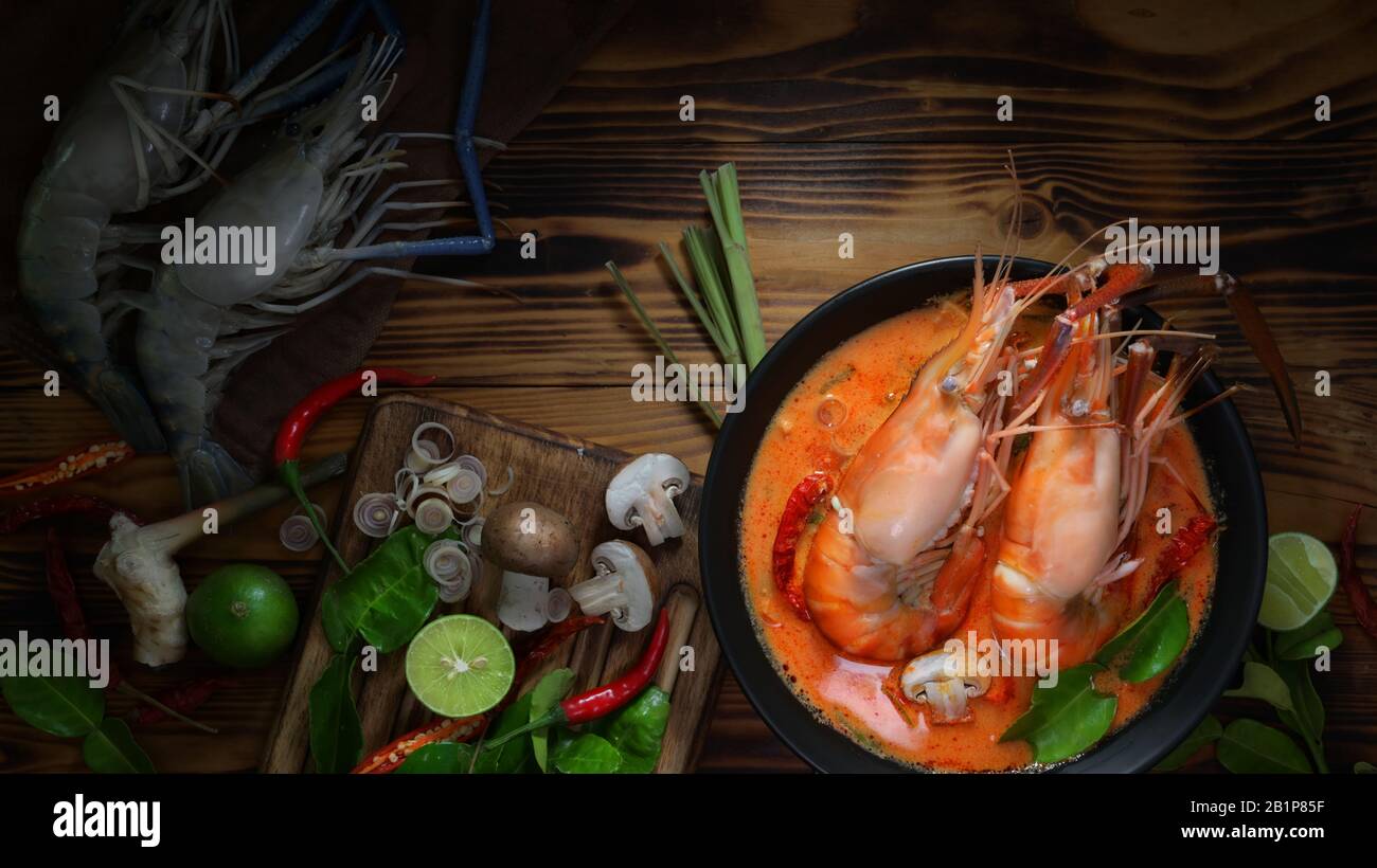 Draufsicht auf Tom Yum goong, thailändische traditionelle Küche mit Garnelen, heiße würzige Suppe in schwarzer Schüssel und thailändische Kräuterzutaten auf Holztisch Stockfoto