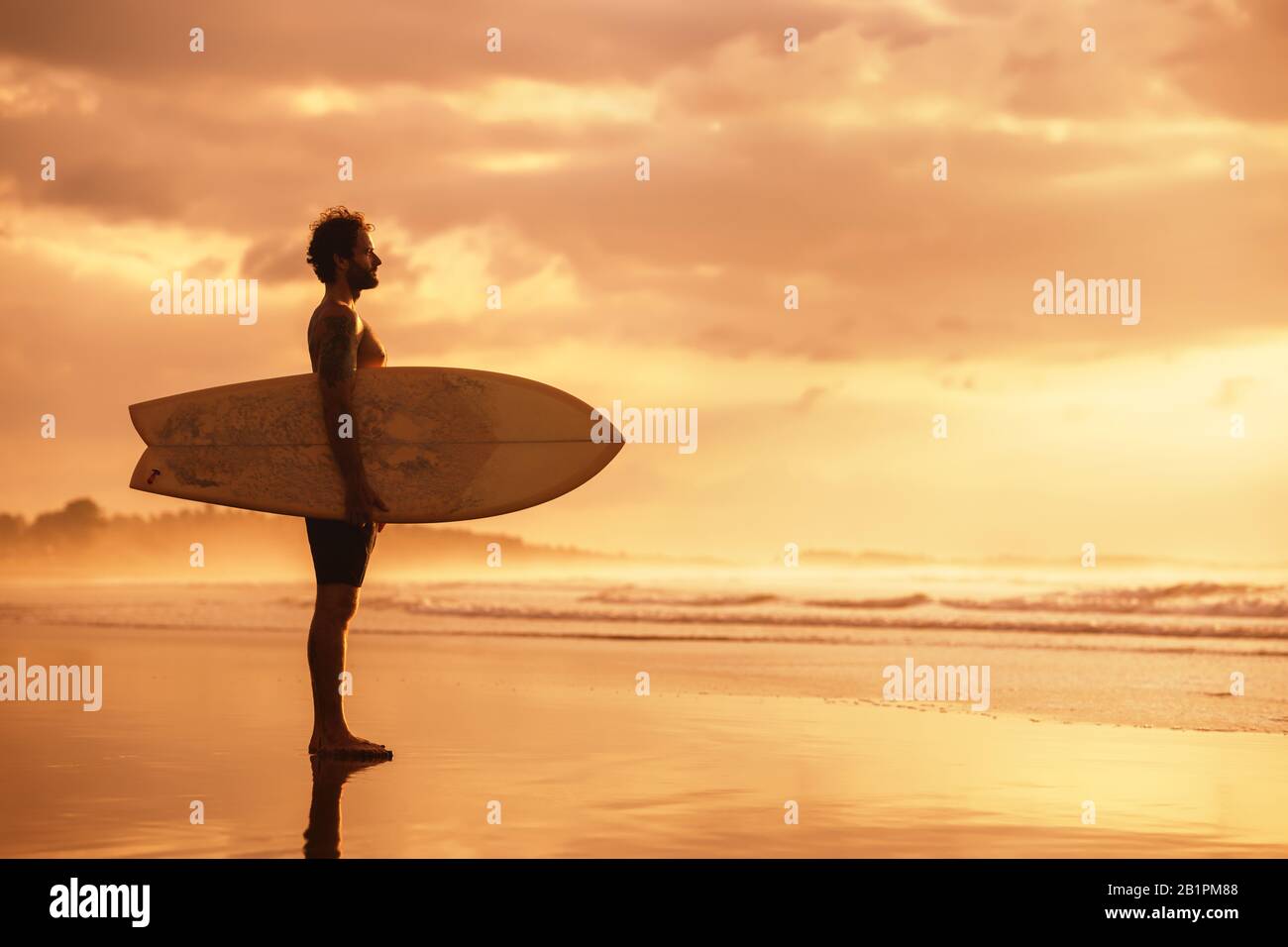 Attraktiver sportlicher Real-Surfer steht mit Surfbrett am Meeresstrand bei Sonnenuntergang Stockfoto