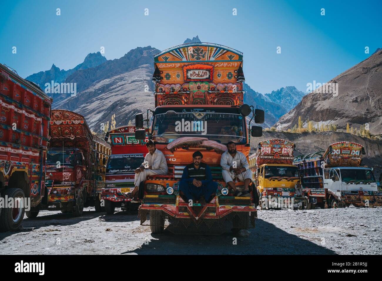 Sust, Pakistan - Oktober 2019: Drei Fahrer sitzen auf einem leuchtend orangefarbenen Truck, der mit bunten Mustern auf einem Bergparkplatz mit anderem TR dekoriert ist Stockfoto