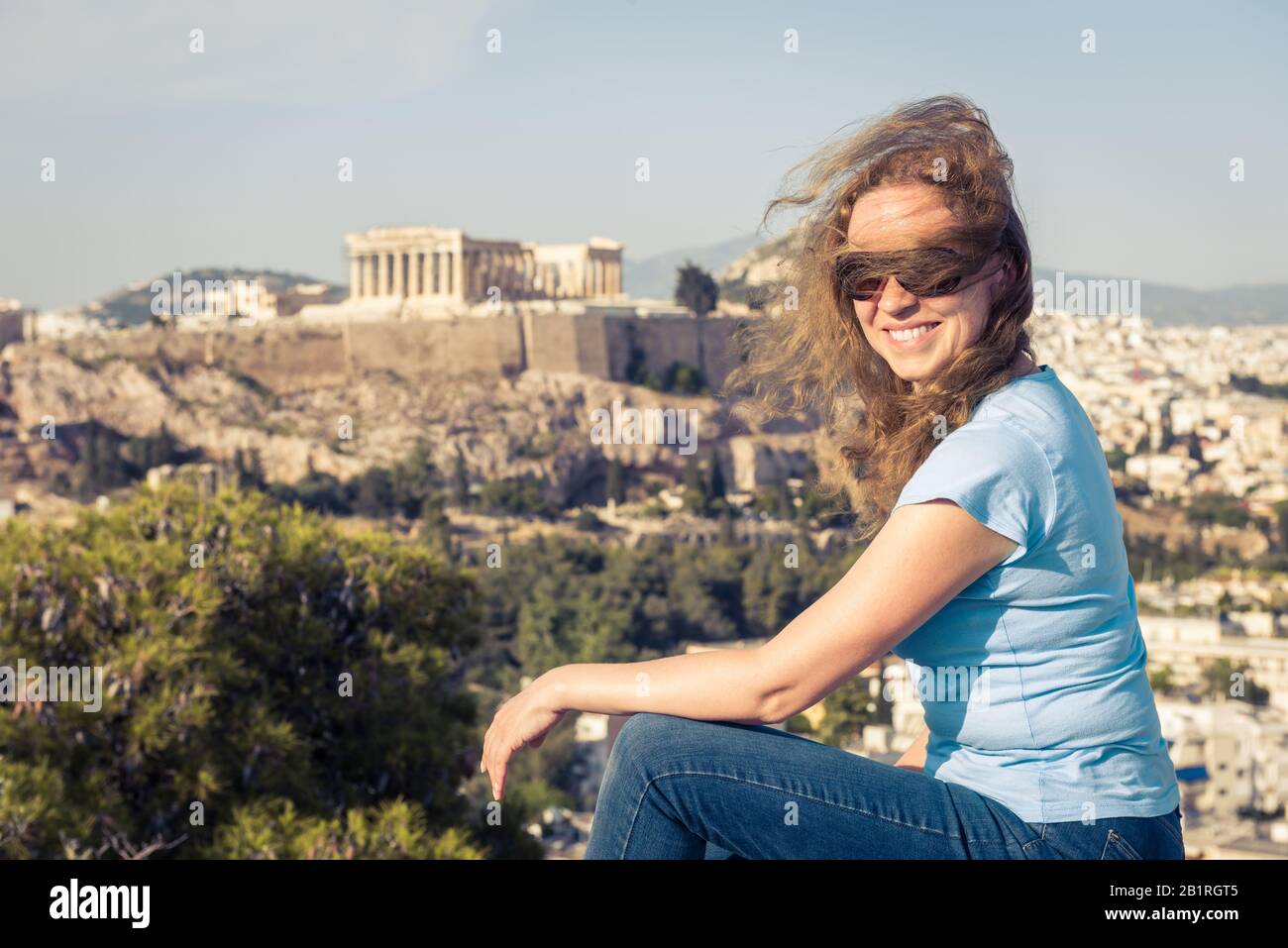 Junge hübsche Frau sitzt auf einem Hügel in Athen, Griechenland. Schöne Erwachsene Mädchen Touristen lächeln mit Blick auf das Stadtzentrum von Athen. Attraktive glückliche Person agai Stockfoto