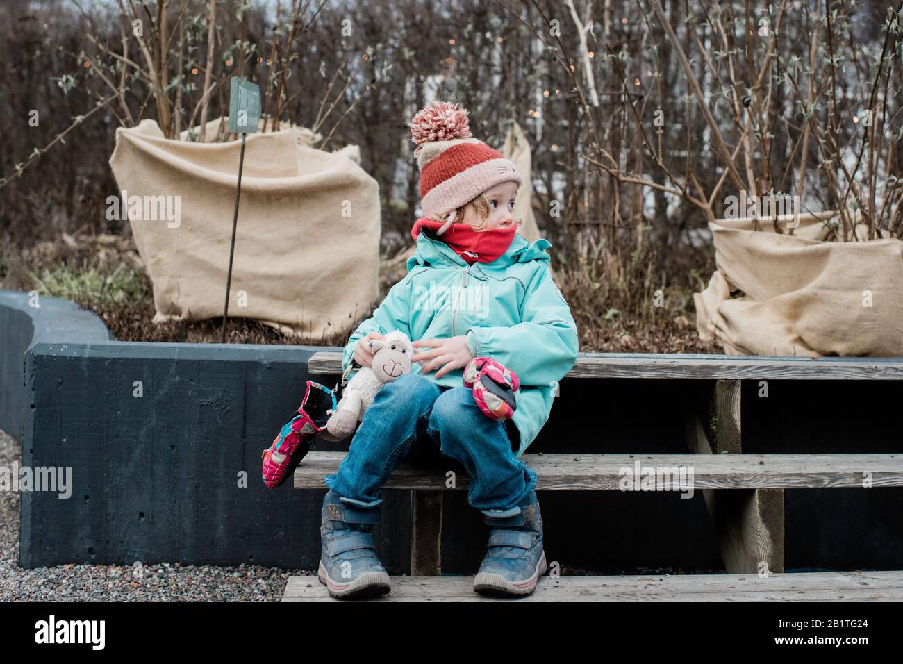 Junges Mädchen eingewickelt warm sitzen mit ihren Spielsachen in einem Garten im Winter Stockfoto