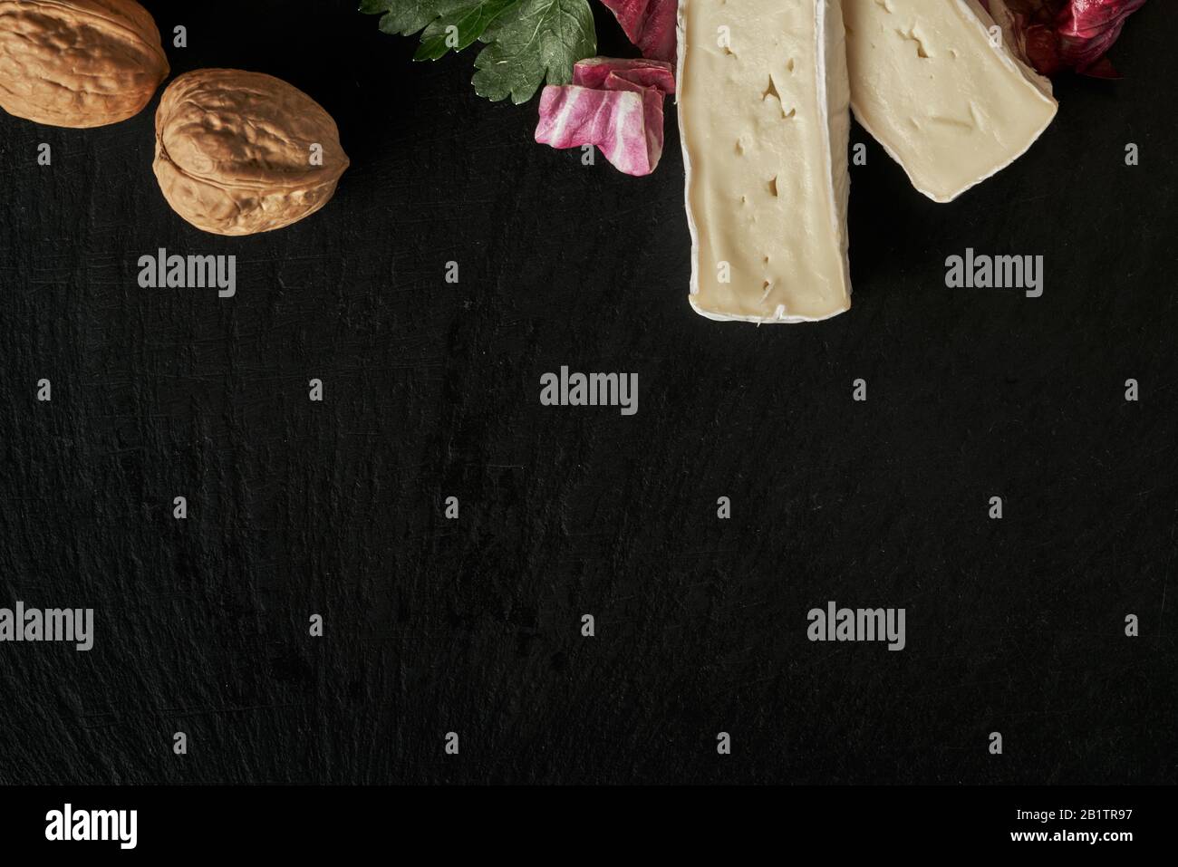 Käsedelikatessen auf schwarzer Steinpultoberfläche. Camembert oder brie kreisen in braunem Kraftpapier, das mit Basilikum und Kirschtomat verziert ist Stockfoto