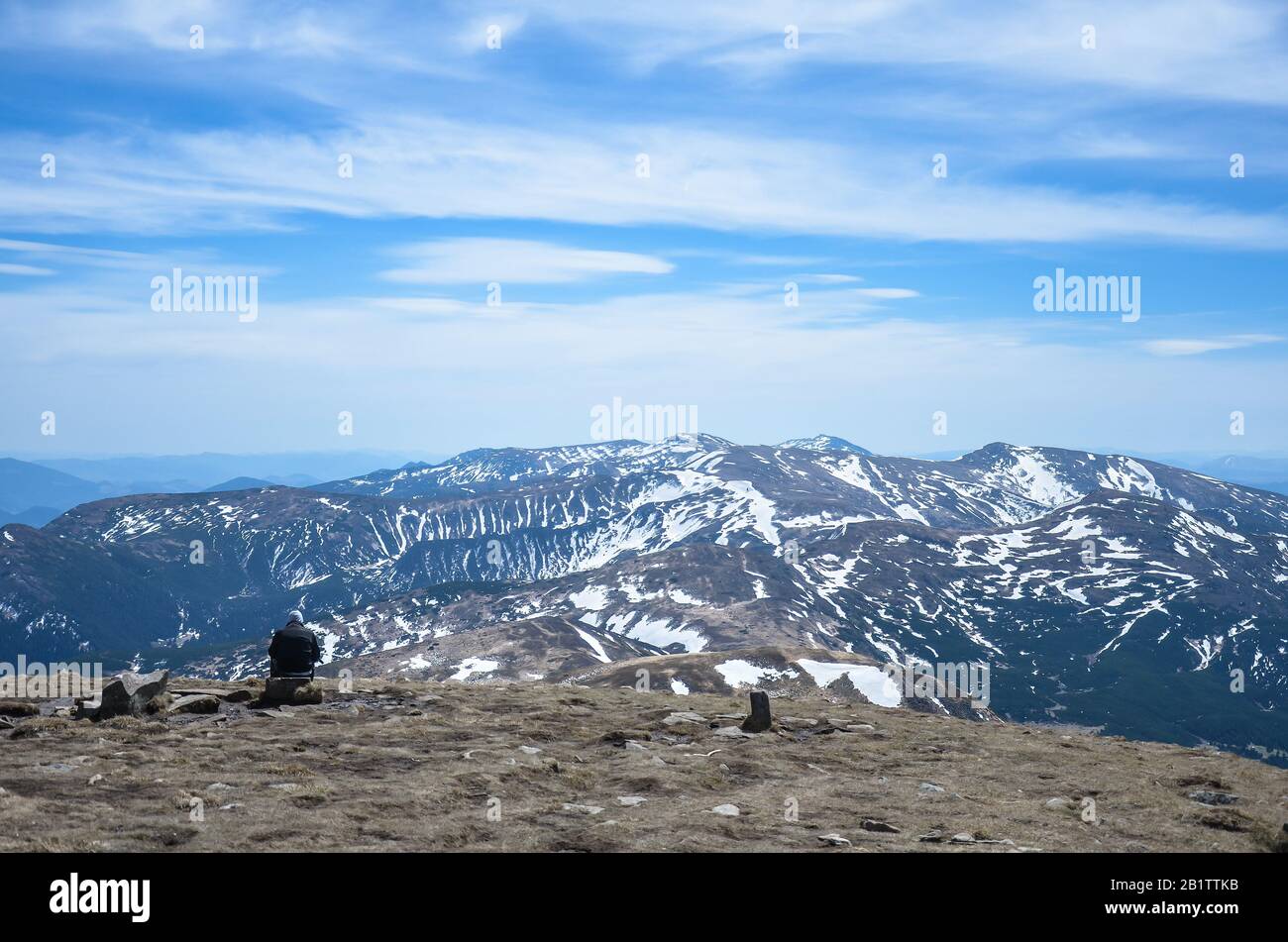 Man sitzt allein und betrachtet die schönen schneereichen Berggipfel am sonnigen Tag, Karpatengebirge, Ukraine. Man Traveler sitzt auf Felsen Stockfoto