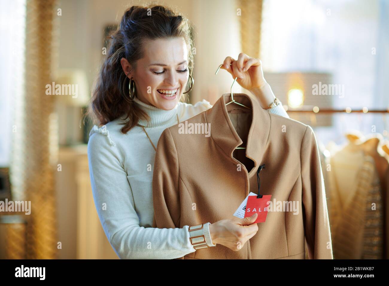 Lächelnde, elegante Frau in weißem Pullover und Rock mit beigefarbenem Mantel mit Preisschild im modernen Modegeschäft. Stockfoto