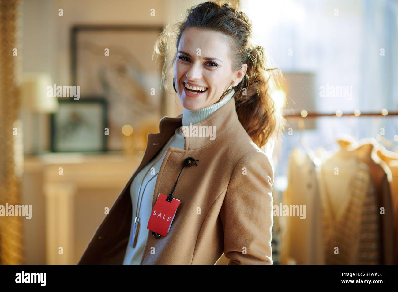 Lächelnde elegante Frau in weißem Pullover und Rock mit beigefarbenem Mantel mit rotem Preisschild in moderner Modeboutique. Stockfoto