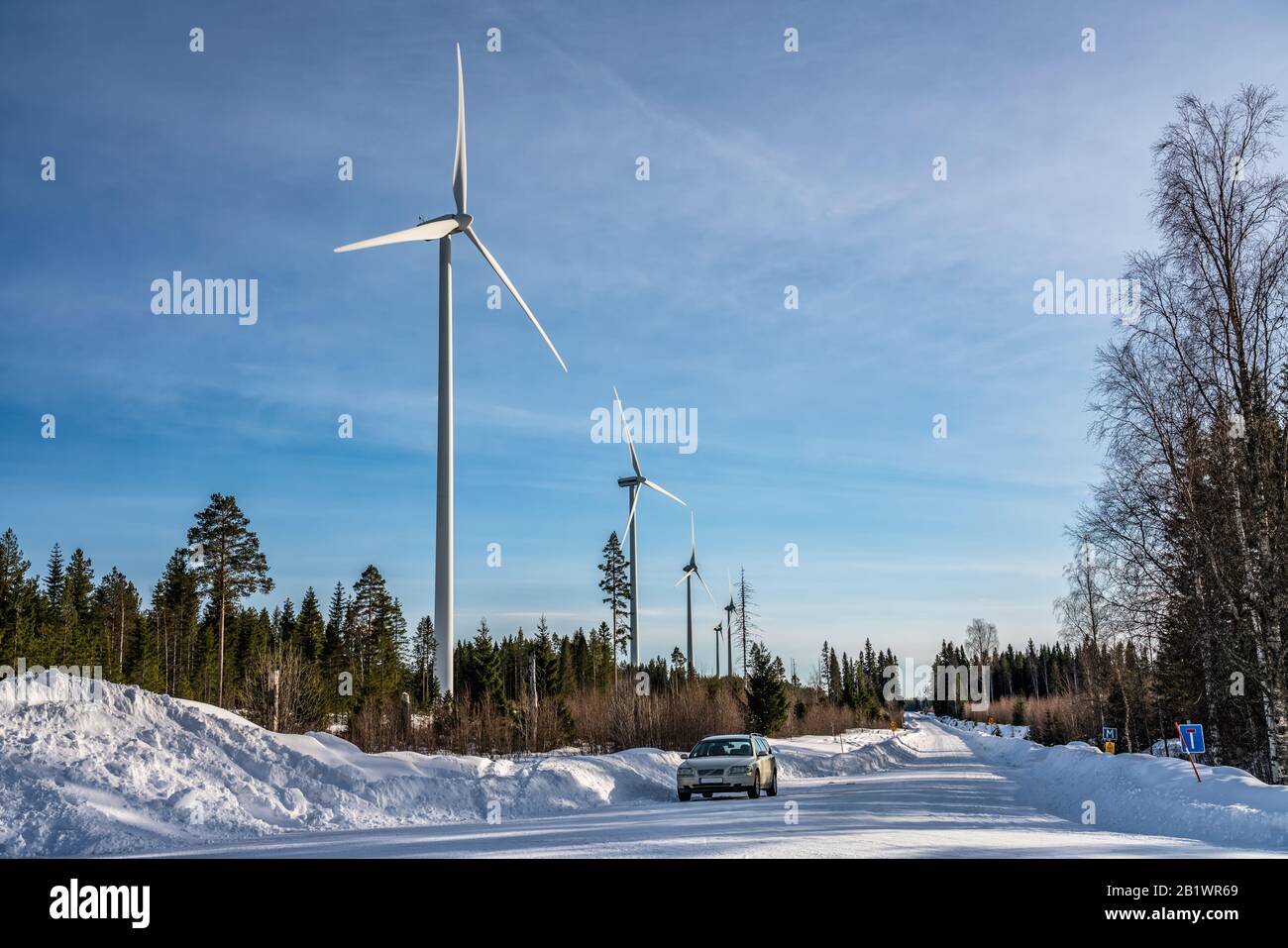 Foto von Windmühlenfarm und Auto auf geraden Straße im Winterwald, blauer Himmel, sonnig, Schweden, umweltfreundliche grüne Energie Stockfoto