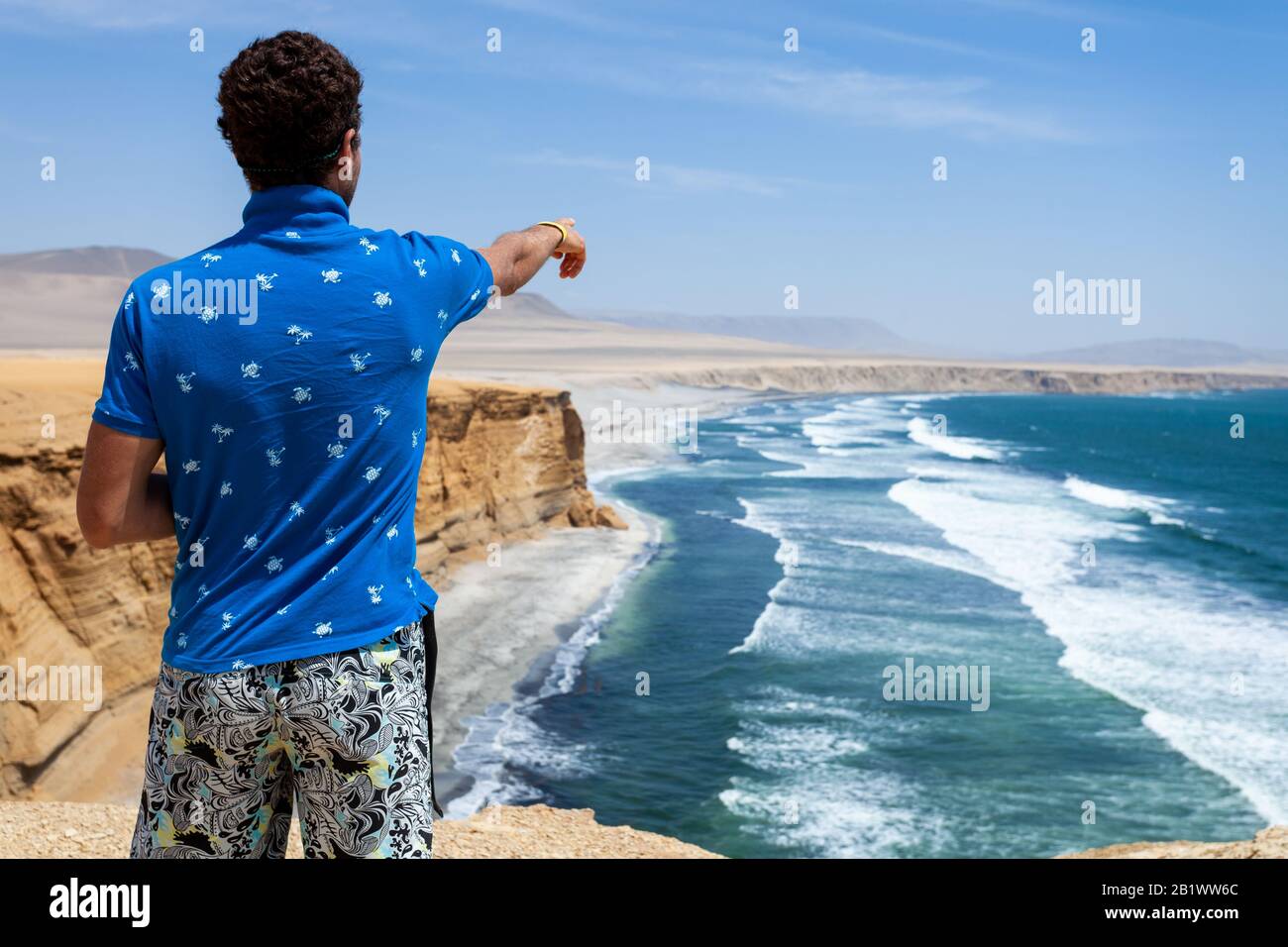 Ein Mann in einem blauen T-Shirt steht auf einer Klippe und zeigt zum Meer Stockfoto