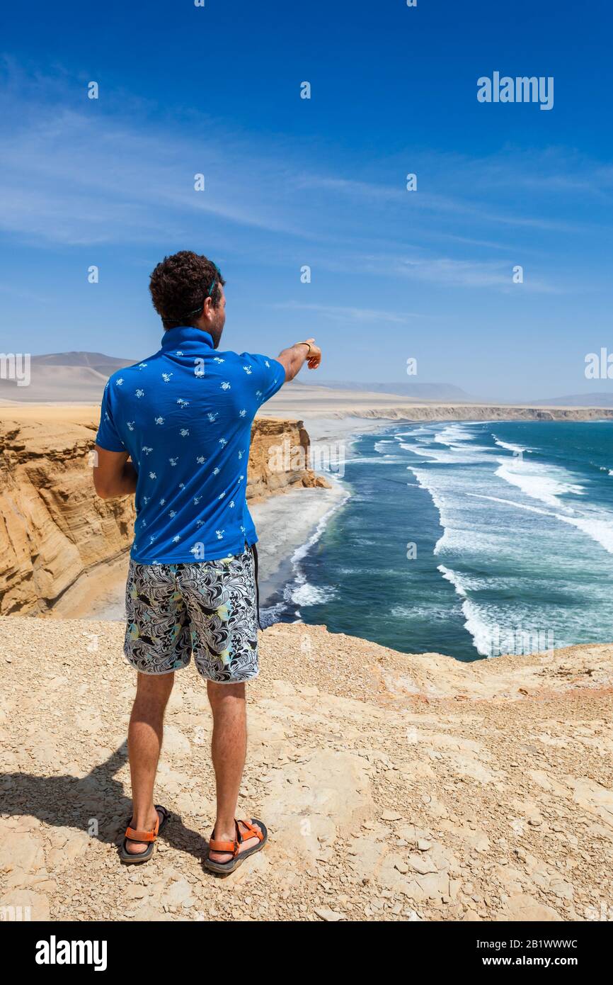 Ein Mann in einem blauen T-Shirt steht auf einer Klippe und zeigt zum Meer. Vertikale Ausrichtung. Stockfoto