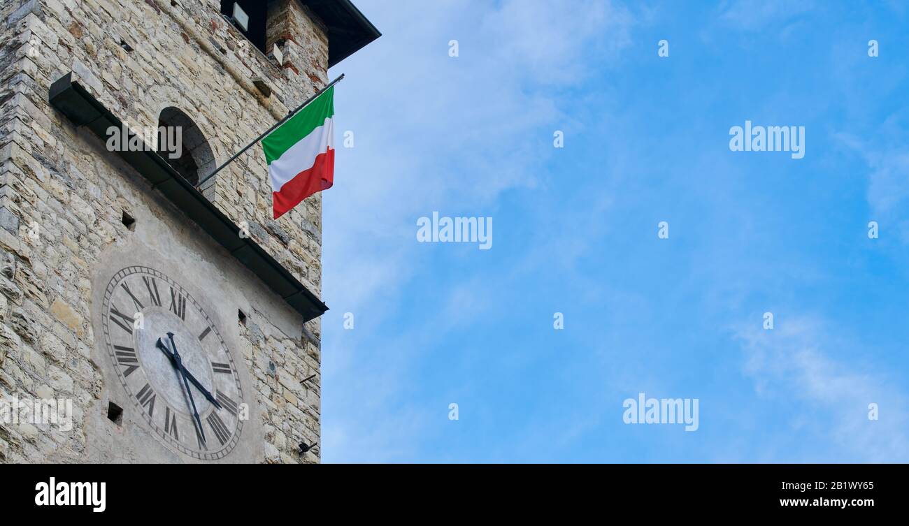 Iseo Lake, Pisogne, Brescia, Italien - 26. Dezember 2019: Alter Steinuhrturm in pisogne mit italienischer Flagge in einem blauen Himmelstag Stockfoto