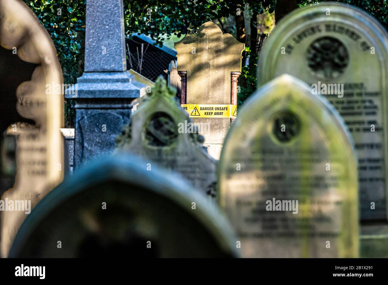 Ein unsicherer Grabstein mit gelbem Warnband. Das Band ist gelb, schwarzer Text und gibt an, dass es sich um ein Ausrufezeichen innerhalb eines schwarzen Dreiecks handelt. Umgeben von zahlreichen alten Kopfsteinen. Stockfoto