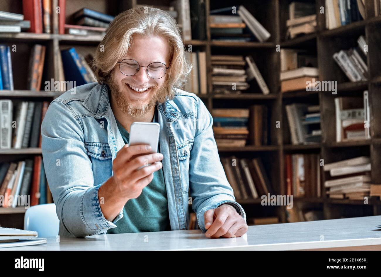 Der kleine bärtige Bücherei-Schalter hält modernes Smartphone am Bildschirm lachen. Stockfoto