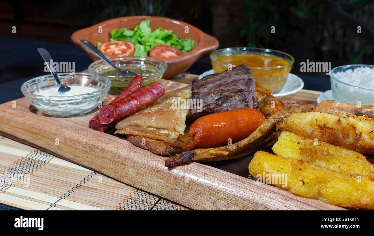 Großes Holztablett mit gegrilltem Fleisch und Würstchen, begleitet von Soßen, gebratenem Maniok, einem Teller mit Reis und Tomaten und Salat auf einem schwarzen Tisch Stockfoto