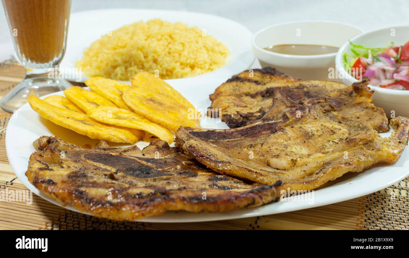 Weiße Platte mit Schweinekopf, begleitet von gebratener Banane, Salat und gelbem Reis mit einer Glaskasse mit Limonade auf weißem Hintergrund Stockfoto