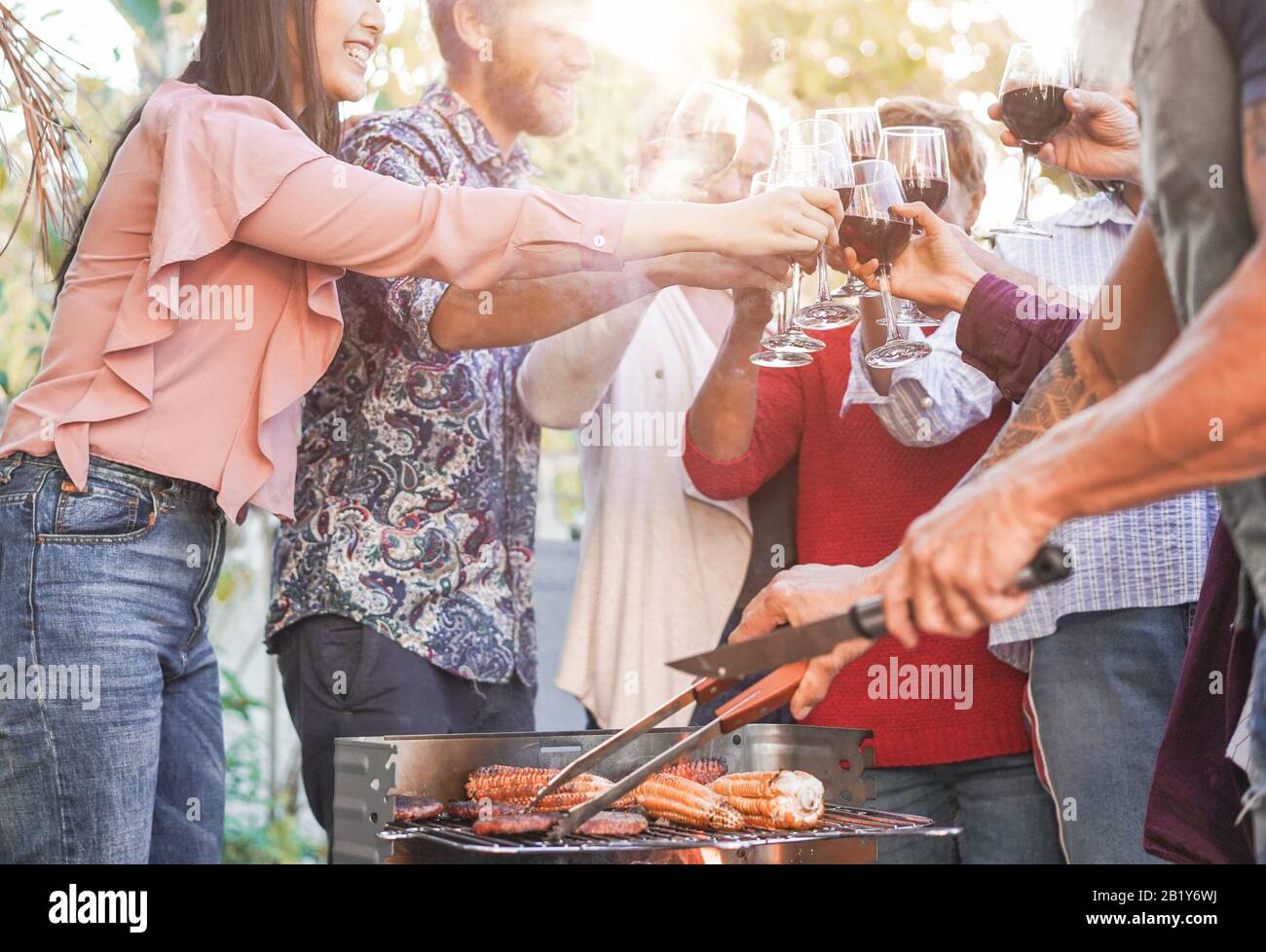 Familie, die Burger, Corns und Jubel mit Rotwein beim grillmenü im Freien zubereitet - Verschiedene Altersgruppen, die Spaß am Grillabendessen haben - Sommerlifestyl Stockfoto