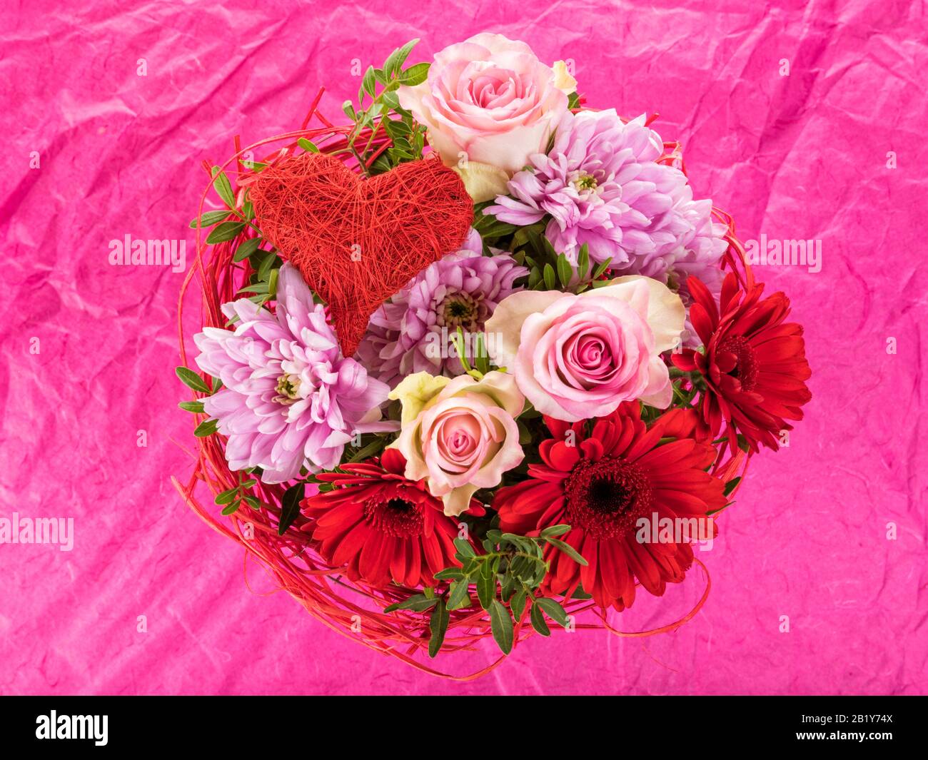 Roter und rosafarbener Blumenstrauß mit Rosen, gerberblüten, Dahlien und herzförmigem Schmuck auf rosa Hintergrund Stockfoto