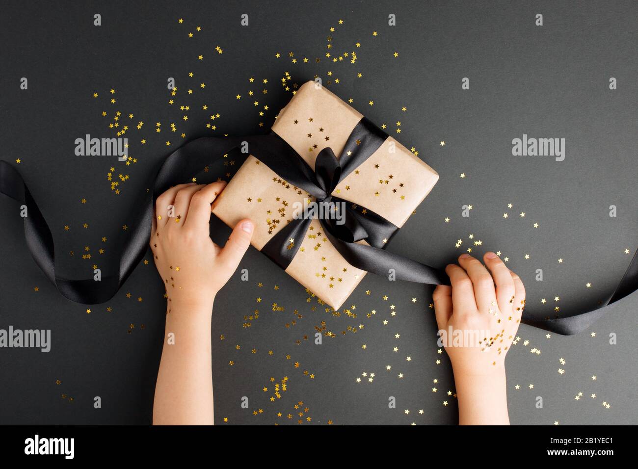 Geschenkschachtel mit Kunsthandwerk auf dunklem Hintergrund, mit Schleife und Glitter, Kinderhänden für eine Veranstaltungsatmosphäre. Zum Geburtstag, Jubiläumsgeschenke, Geschenk Stockfoto