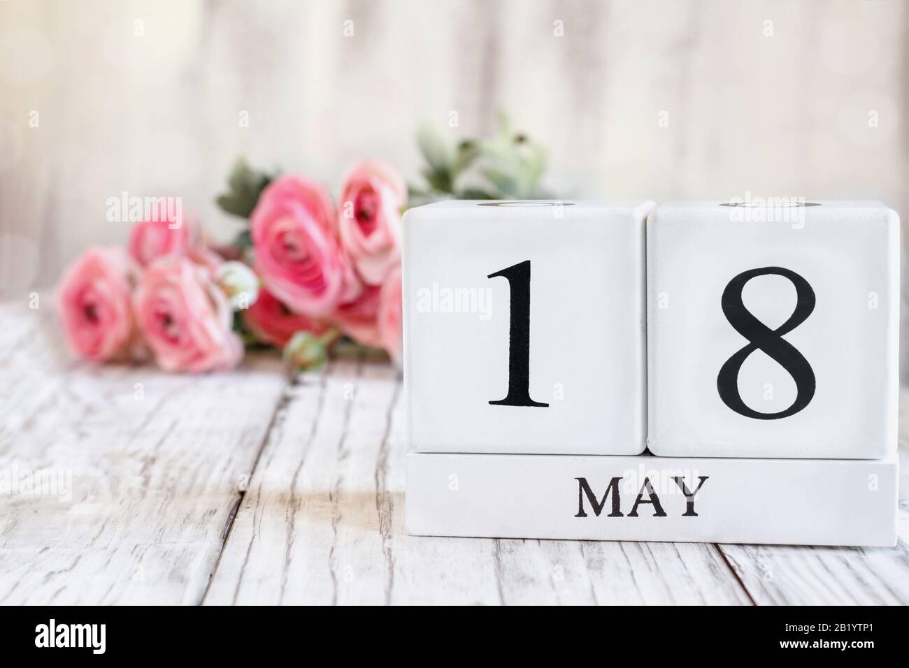 Weiße Holzkalendarkaden mit dem Datum 18. Mai. Selektiver Fokus mit pinkfarbenem Ranunculus im Hintergrund über einem Holztisch. Stockfoto