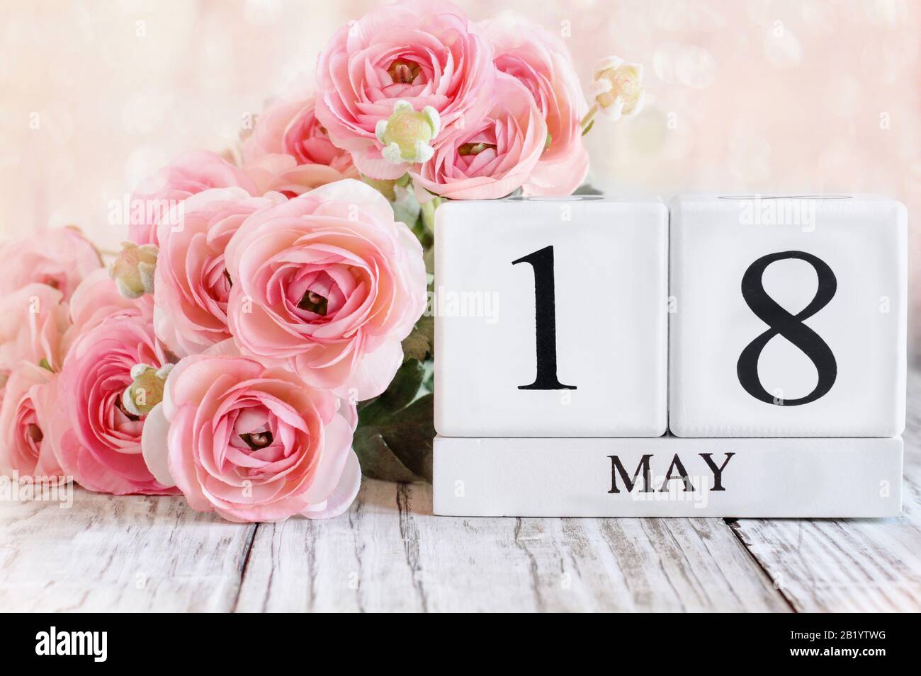 Weiße Holzkalendare mit dem Datum 18. Mai und rosafarbene Ranunculus Blumen über einem Holztisch. Selektiver Fokus mit verschwommenem Hintergrund. Stockfoto