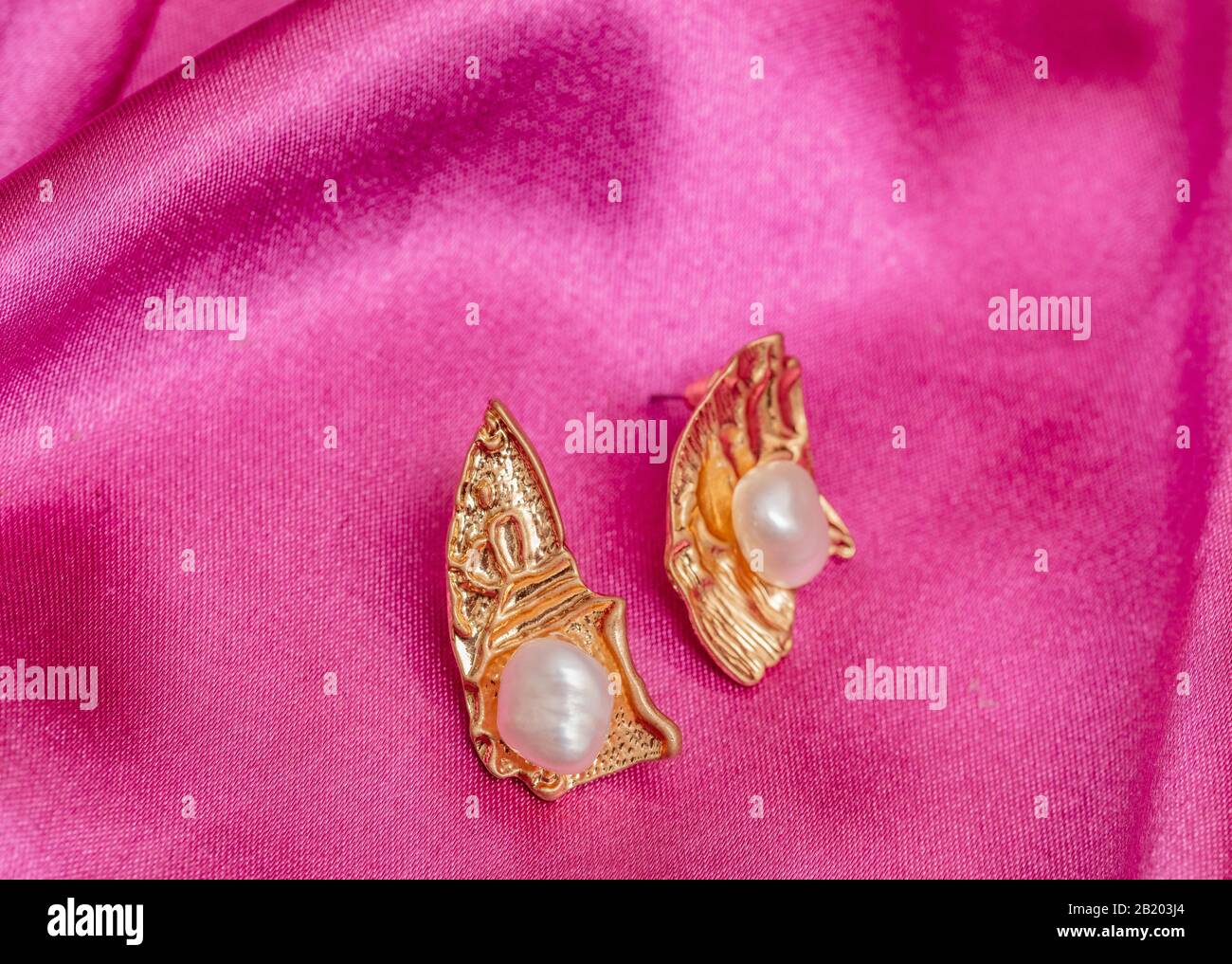 Moderne asymmetrische goldene Ohrringe mit Perlen auf pinkfarbenem Seidenhintergrund Stockfoto