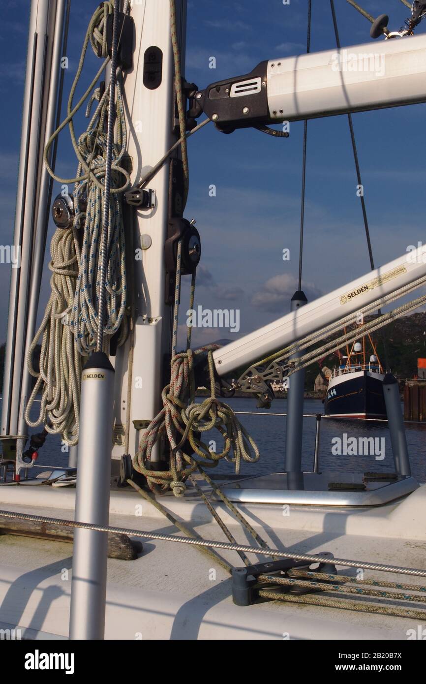 Ein Nahblick auf einen Mast, Segel und Takelage auf einem privaten Segelboot mit Fischerboot und Hafen im Hintergrund Stockfoto