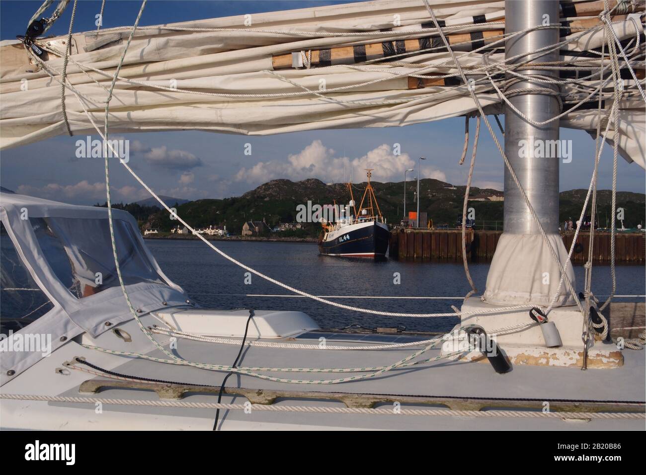 Nahansicht von Mast, Kabine, Segel und Takelage auf einem privaten Segelboot mit Fischerboot und Hafen im Hintergrund Stockfoto