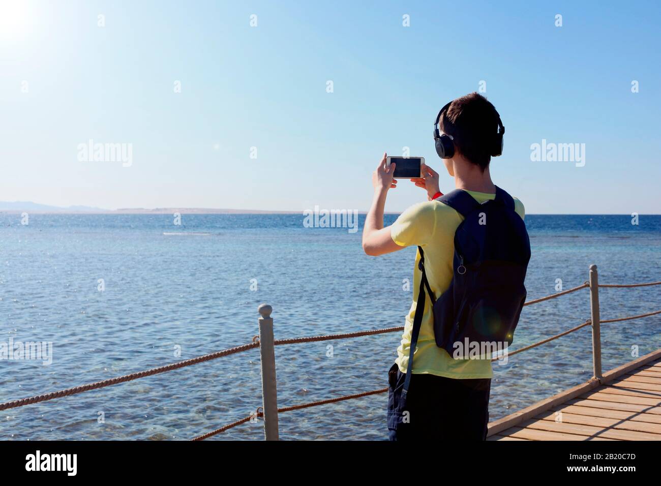 Hiphster Guy mit Kopfhörern, die auf der Digitalkamera eines Mobiltelefons in der Nähe des Meeres Bilder von wunderbarer Landschaft machen, wunderschönes Sonnenlicht Stockfoto