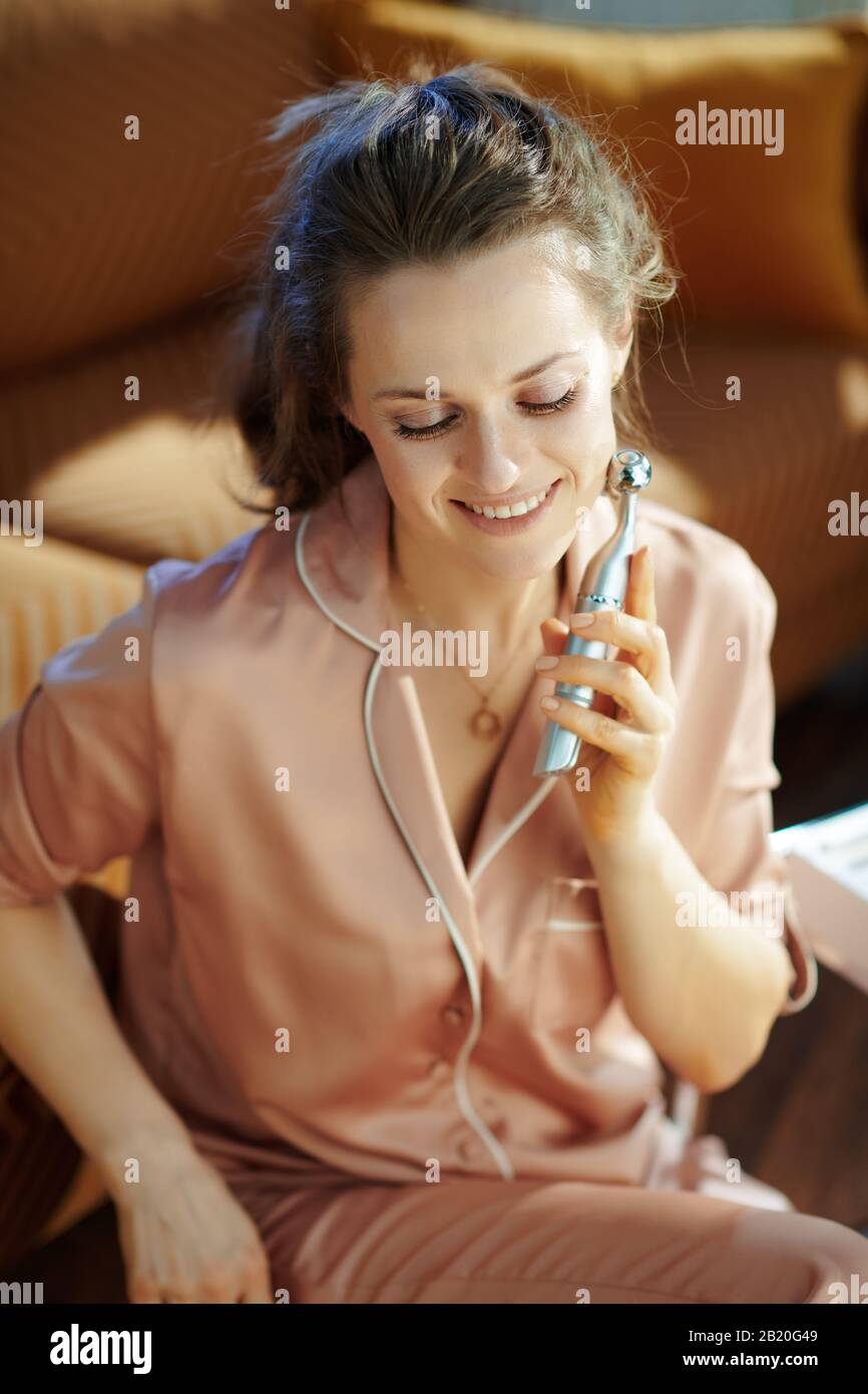Entspannte moderne 40 Jahre alte Hausfrau im Schlafanzug im modernen Wohnzimmer am sonnigen Wintertag mit Gesichtsauszug Anti-Aging-Elektro-Massagegerät. Stockfoto