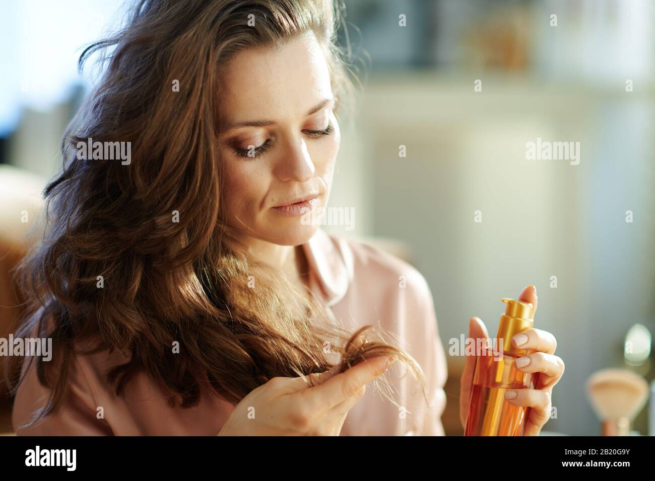 Besorgt über die moderne 40 Jahre alte Frau im Schlafanzug mit langen, welligen Haaren, die die Haarölflasche halten und Haarspitzen im modernen Wohnzimmer i prüfen Stockfoto