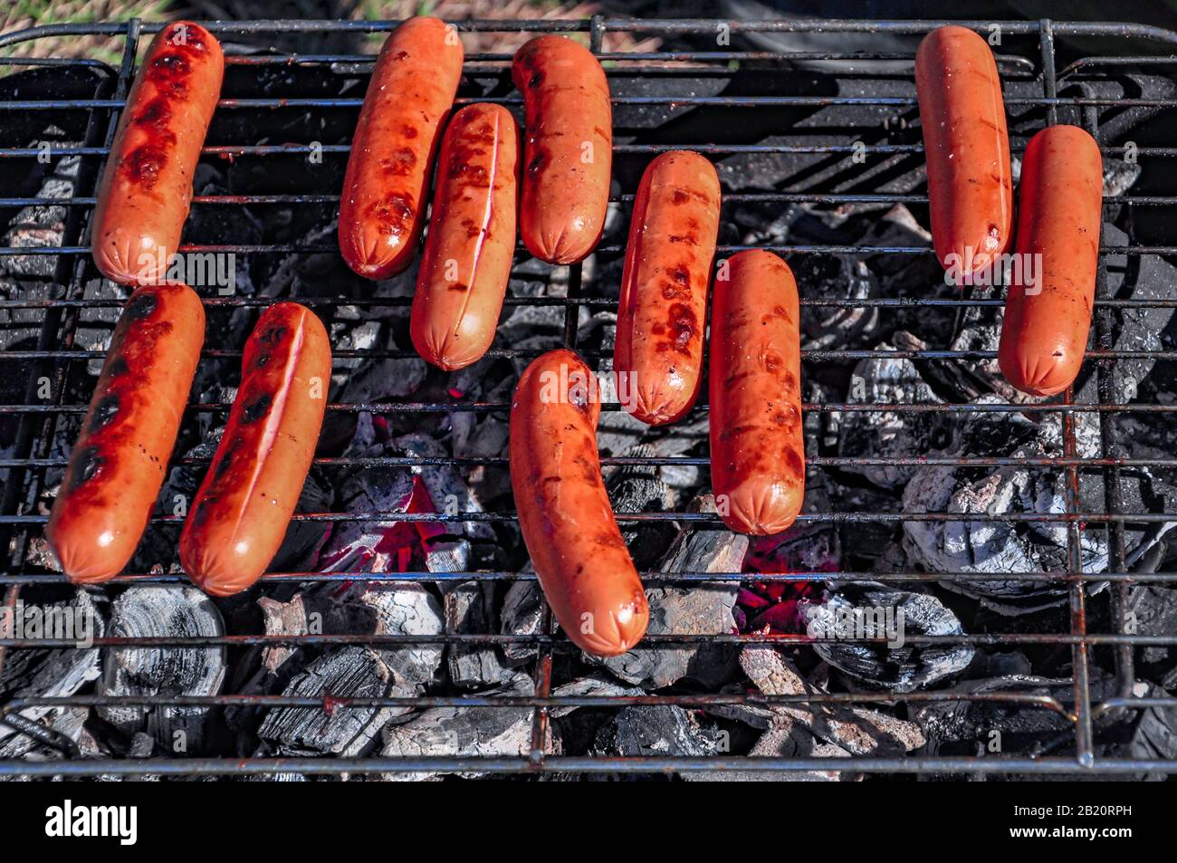 Auf dem Gras stehend, ein kleiner tragbarer Picknickgrill mit brennenden Kohlen und Würstchen, die auf dem Grill braten und von den Strahlen der Einstellung su hell erleuchtet werden Stockfoto