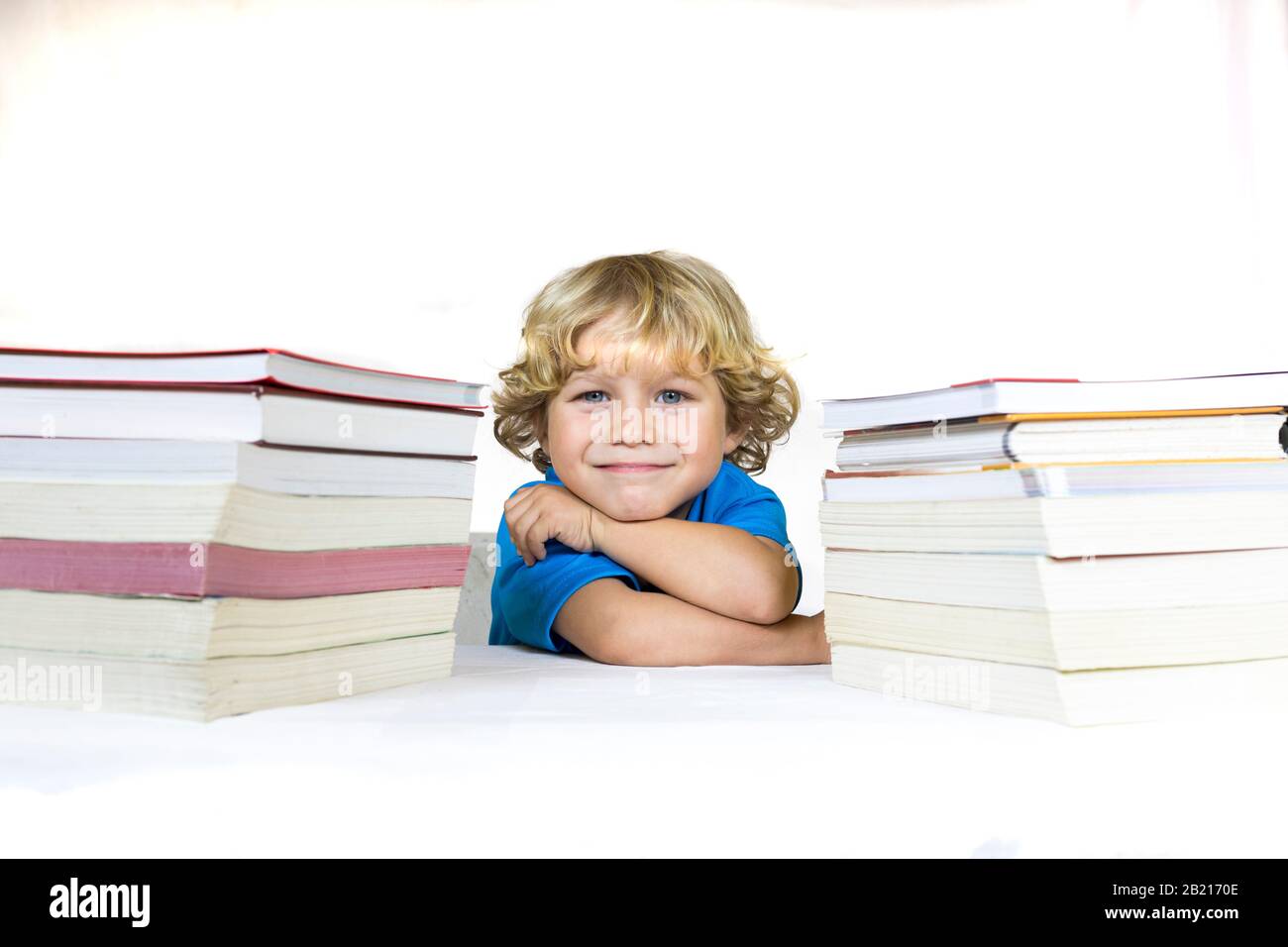 Kleines glückliches Kind zwischen 4 und 5 Jahren sitzt an einem Schreibtisch und studiert mit Lehrbüchern auf seinem Tisch und mit weißem Hintergrund Stockfoto