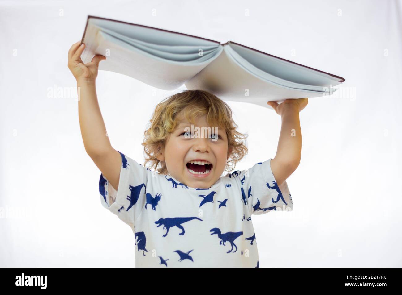 Kleiner Junge zwischen 4 und 5 Jahren, der ein Buch über den Kopf hält und dabei vor weißem Hintergrund lächelt Stockfoto