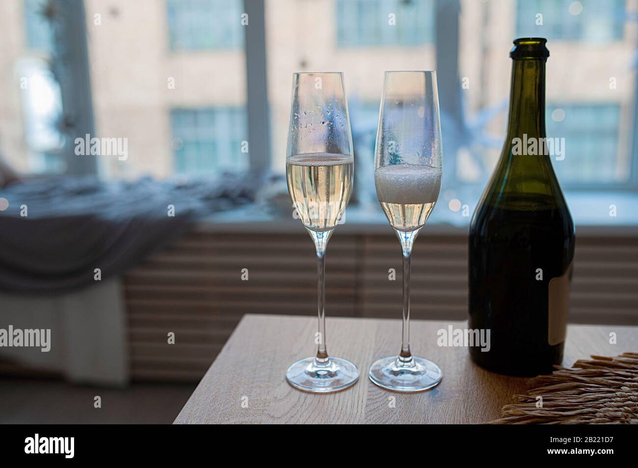 Zwei Perlmuttgläser mit Champagner, Blasen und Schaum neben einer dunklen Glasflasche gegen ein Fenster in Blautönen Stockfoto