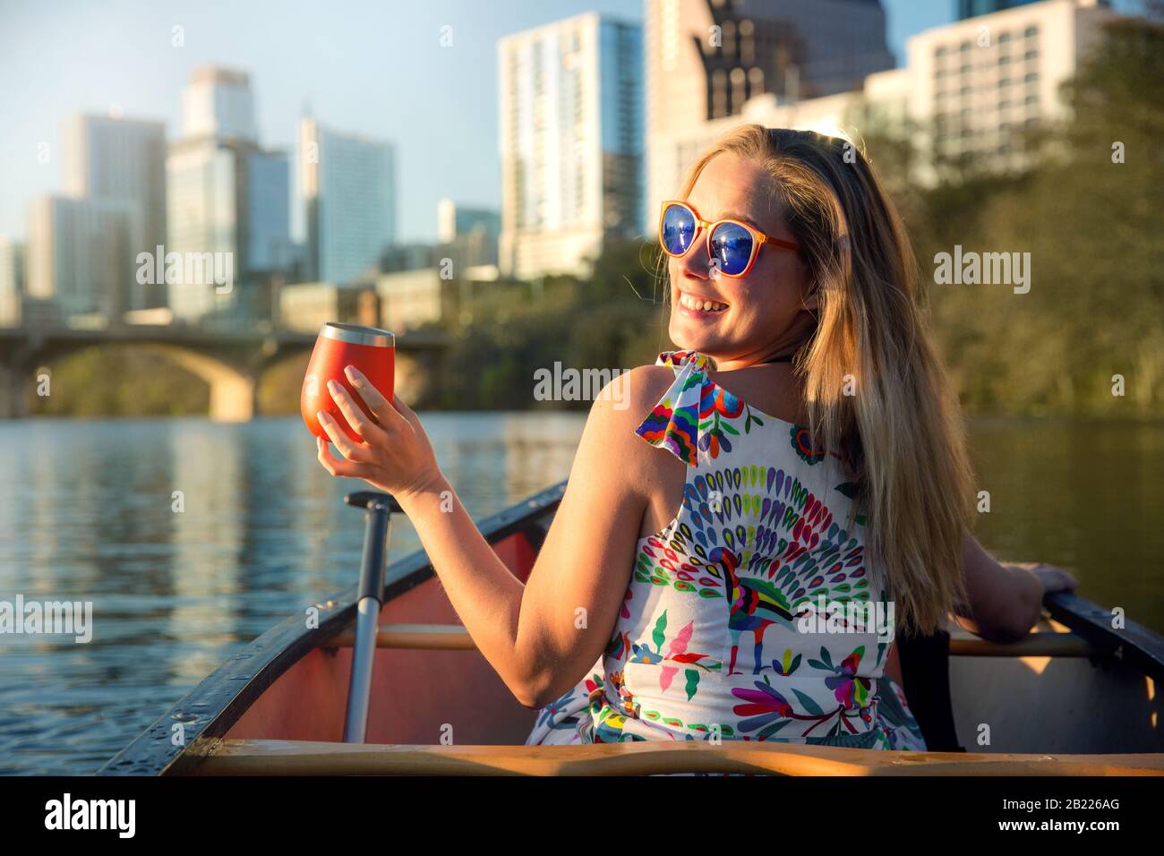 Lustige junge Frau in einer Sonnenbrille lächelt, genießt das Leben und macht Spaß mit dem Boot auf einem Fluss neben einer Skyline der Stadt Stockfoto
