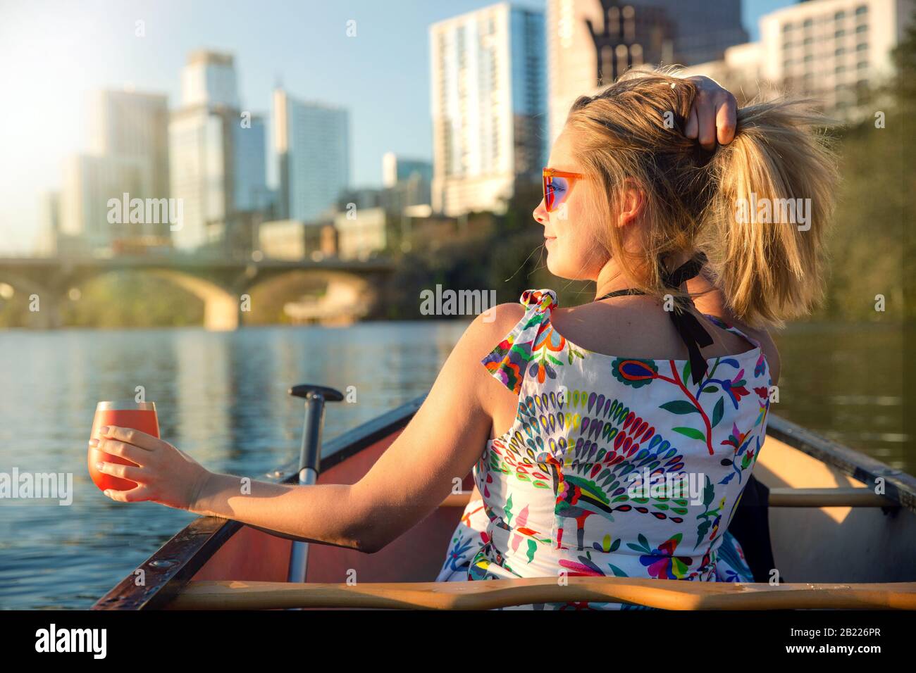 Lustige junge Frau in einer Sonnenbrille lächelt, genießt das Leben und macht Spaß mit dem Boot auf einem Fluss neben einer Skyline der Stadt Stockfoto