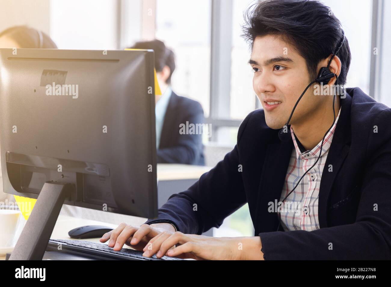 Lächelnder, gutaussehender asiatischer Callcenter-Betreiber verwendet Computer für die Kommunikation mit dem Kunden oder für andere Aufgaben. Stockfoto