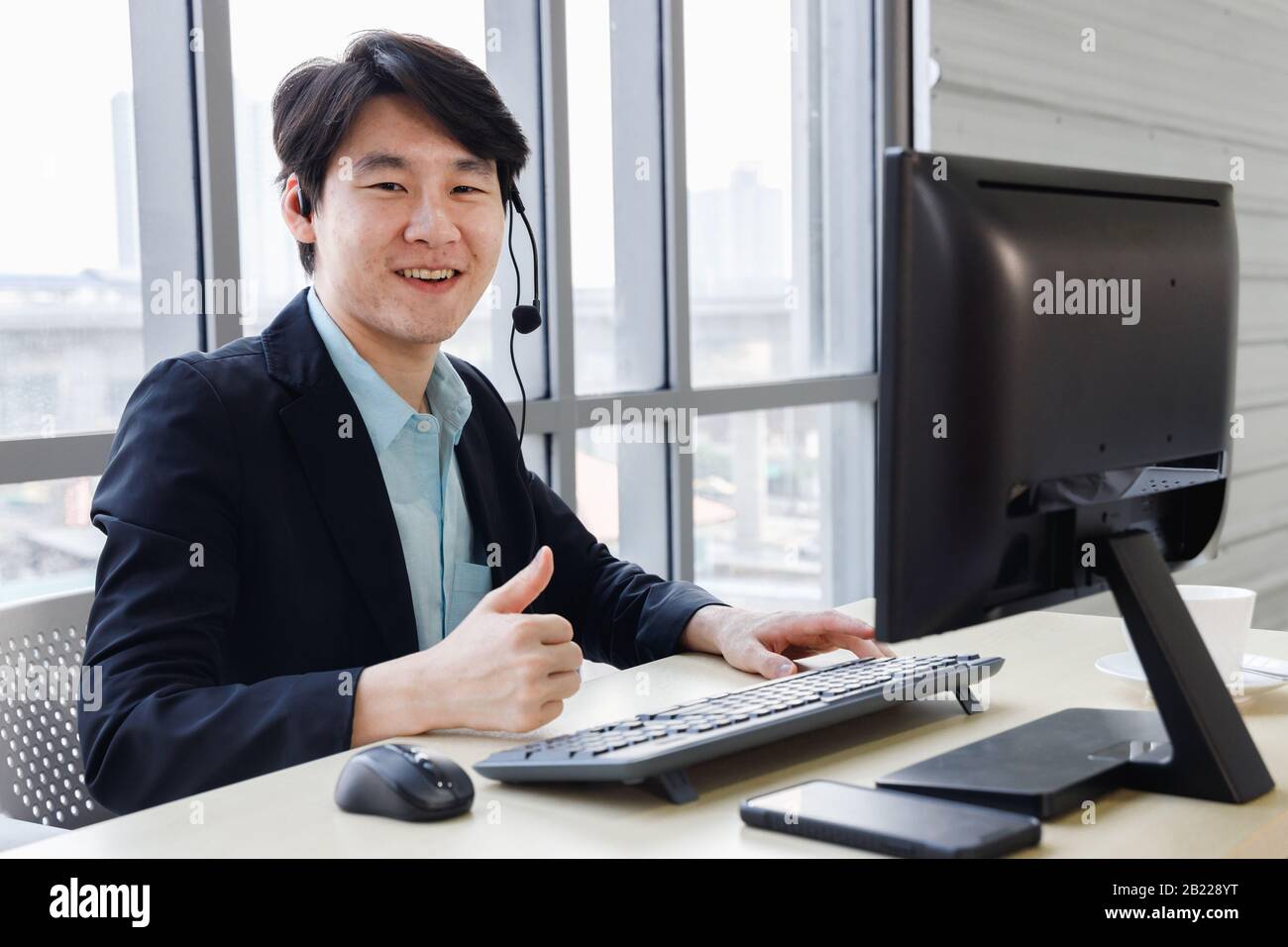 Lächelnder, selbstbewusster asiatischer Callcenter-Betreiber genießt seine Arbeit im Büro und führt eine "Daumen hoch"-Geste durch. Stockfoto