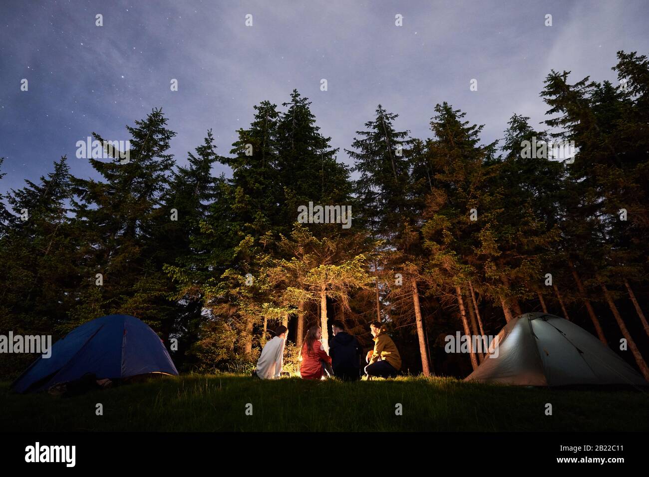 Nachtcamping in der Nähe von Lagerfeuer vor dem Hintergrund eines mächtigen Waldes. Vier Personen sitzen in der Nähe von Feuer- und Touristenzelten. Schöne Fichten unter dem Abendhimmel, auf dem die Sterne auftauchen Stockfoto