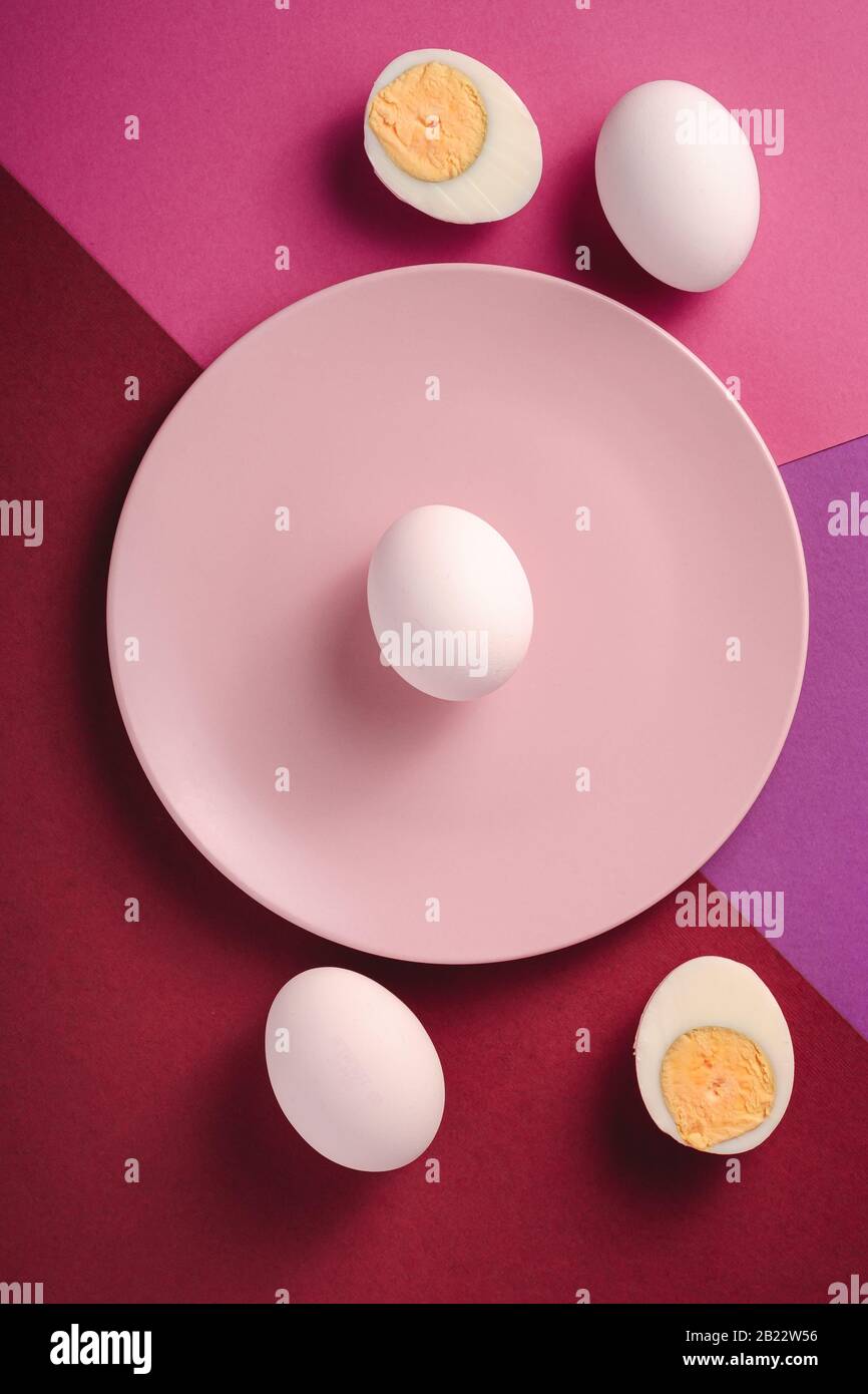 Weißes Ei in pinkfarbener Platte, in der Nähe von geschälten gekochten Eiern in violetten, rosa und roten Farben, minimaler Hintergrund, Draufsicht, glücklicher Ostertag Stockfoto