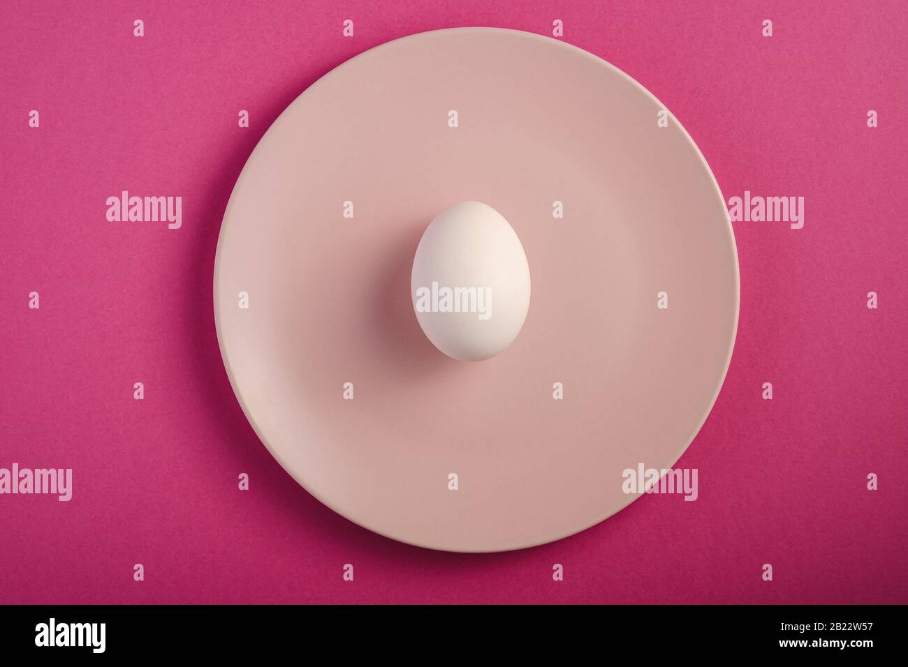 Weißes Ei in pinkfarbener Platte auf purpurfarbenem, minimalem Hintergrund, Draufsicht, fröhlicher Ostertag Stockfoto