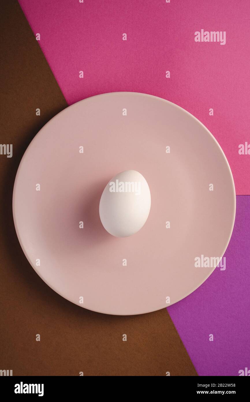 Weißes Ei in pinkfarbener Platte in violetten, rosa und braunen Farben schlichter minimaler Hintergrund, Draufsicht, fröhlicher Ostertag Stockfoto