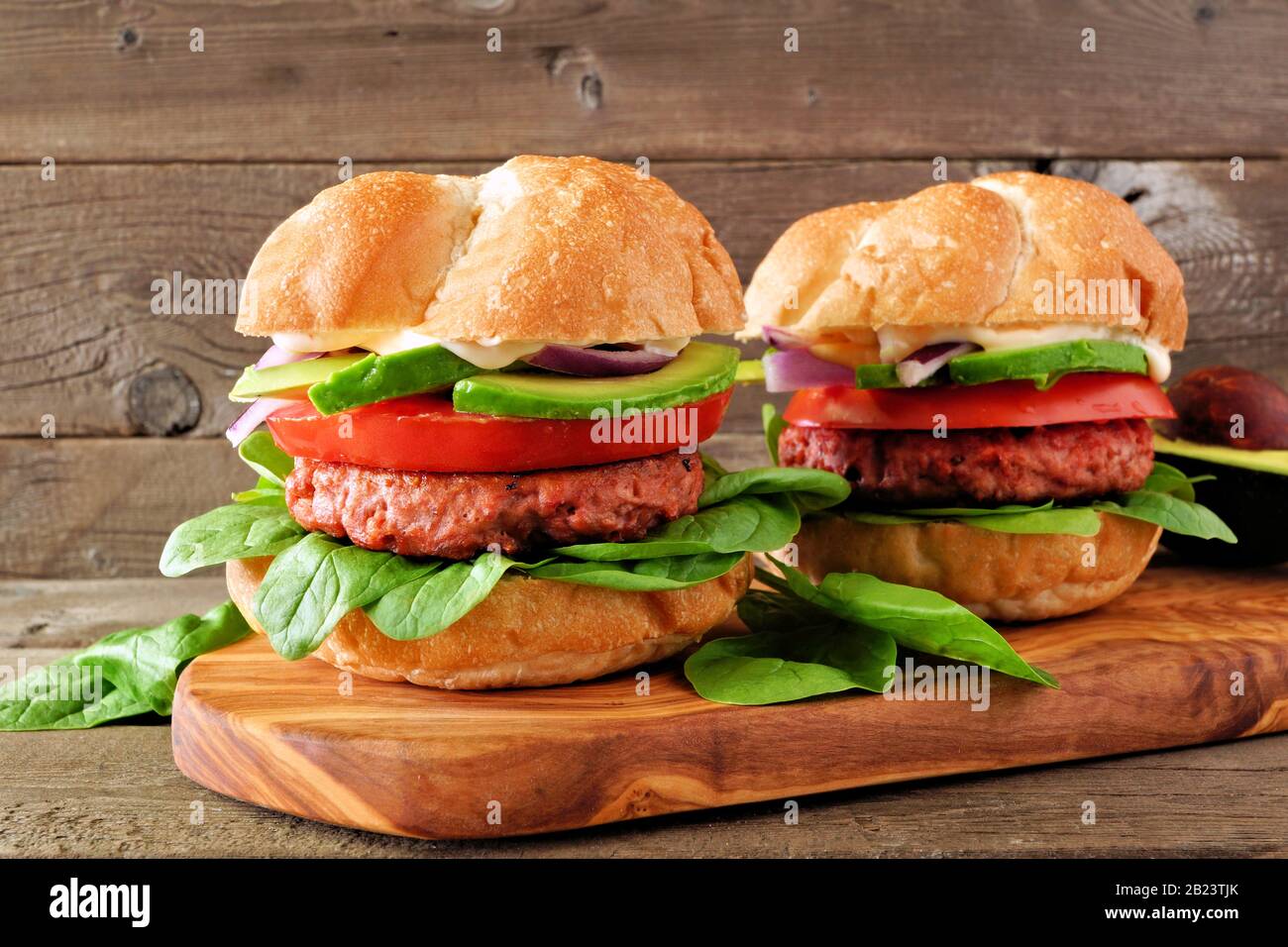 Fleischlose Burger mit Avocado, Tomaten und Spinat auf Pflanzenbatterbasis auf einem Servierbrett vor einem rustikalen Holzhintergrund Stockfoto